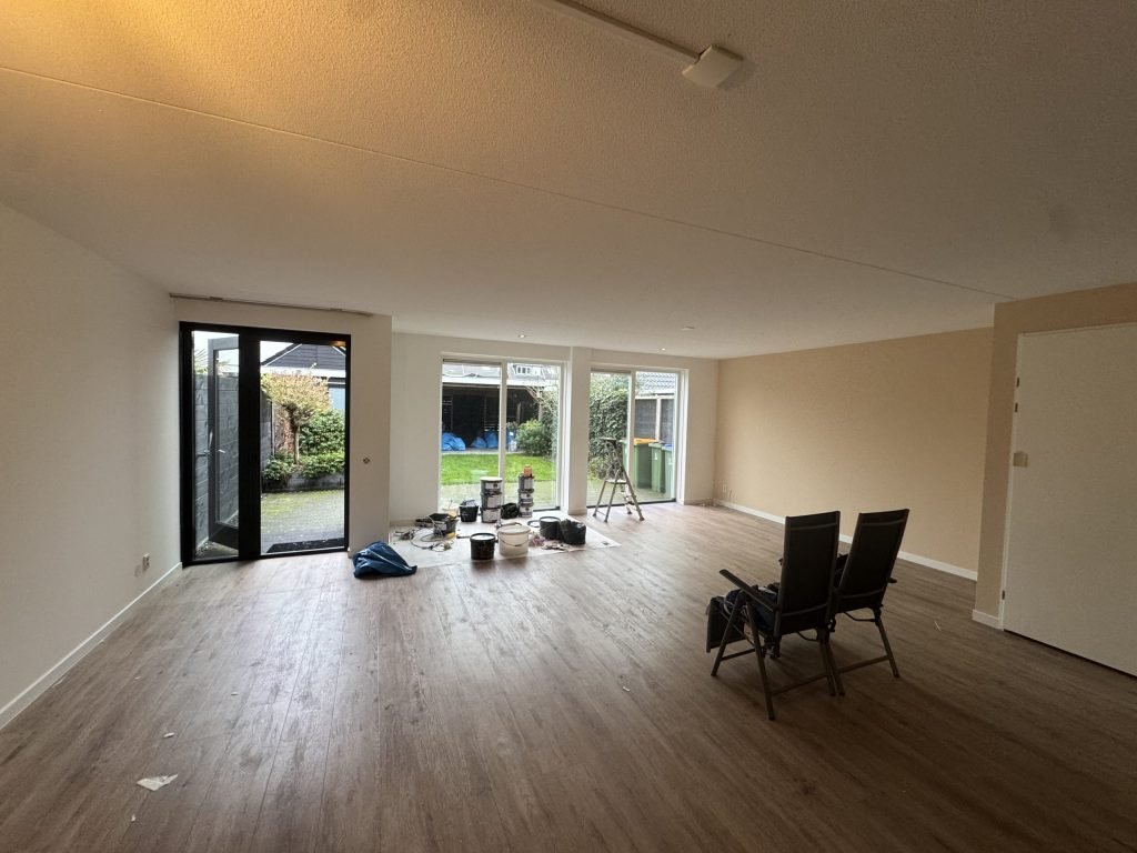Ruime woonkamer van een eengezinshuis met tuindeuren, verfemmers en een trapje op de vloer tijdens het werk